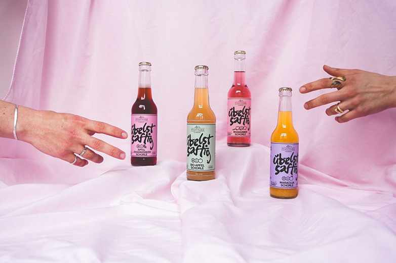 4 Flaschen von der Marke Übelst saftig, die dekorativ auf blass rosa Stoff stehen. Von beiden Seiten greift eine Hand nach je einer Flasche